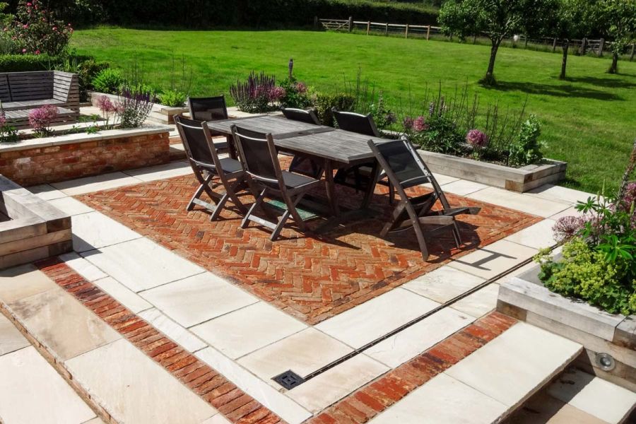 Dining set on clay paving edged with Mint Indian sandstone paving and sleeper-edged beds, next to large lawn with 3 small trees.