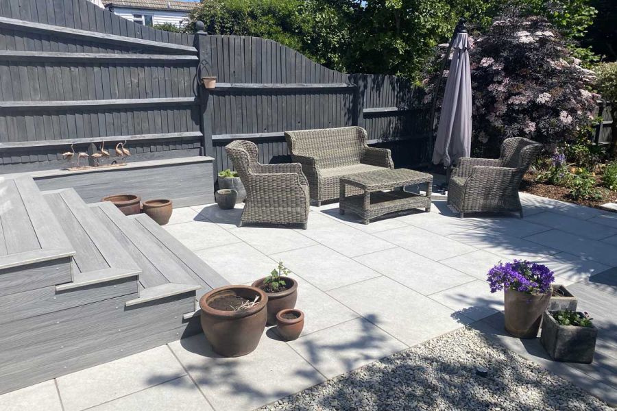 Grey decking steps leading down to Astor Grey Porcelain Paving patio surrounded by black fence, rattan furniture in the corner.