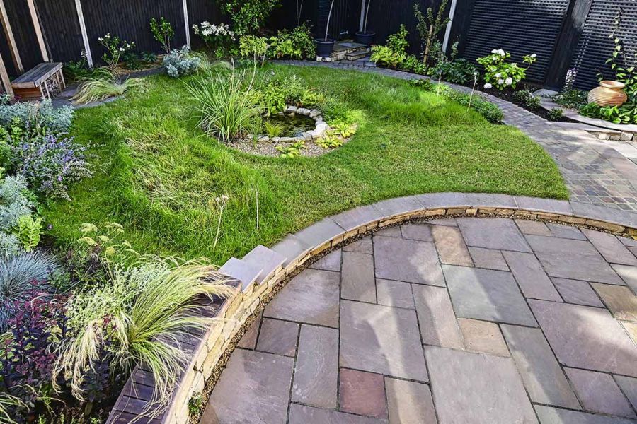Half-moon shaped grassed area edged with Autumn Indian sandstone setts and a low wall encircling a round patio of matching paving.