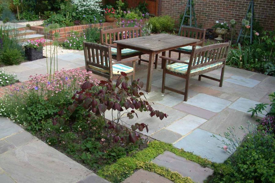 Wooden table with chairs and benches on Autumn Brown Indian sandstone paving with clay paver detail and planted beds. 
