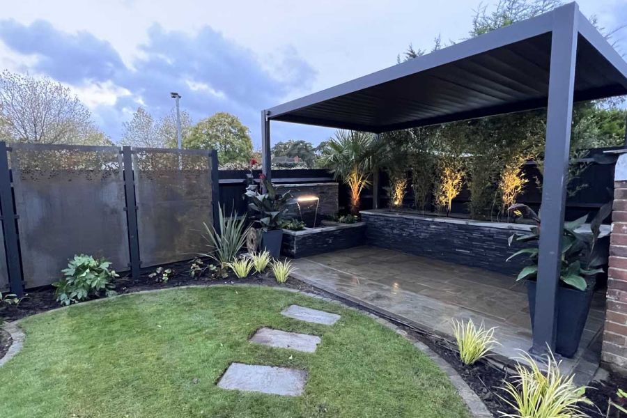 Stepping stones in curved lawn lead to Proteus Grey aluminium pergola over paved area with water feature. Built by Aye Gardening.