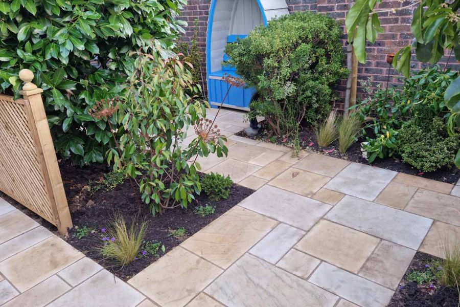 2 oblong beds with shrubs in small area bounded by tall brick wall, paved with Mult Mint sawn sandstone paving. 