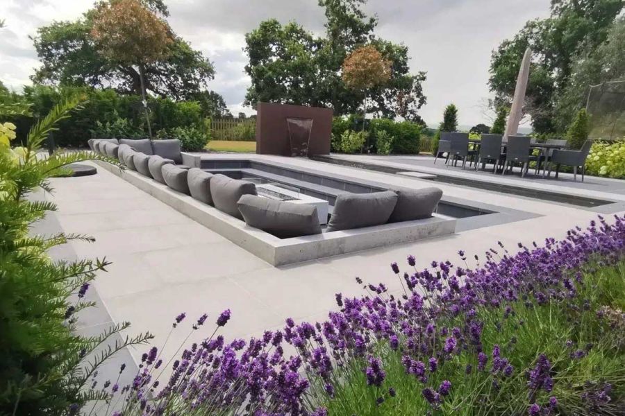 Large rectangular sunken seating area next to long pond with dining area beyond, all in midst of grey Cement porcelain paving.
