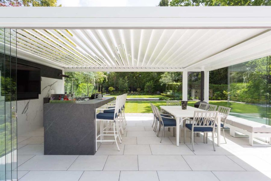 Impressive view of outside patio area paved in Florence White Porcelain Paving with a bar facing a tv and a dining area, under a large white pergola.