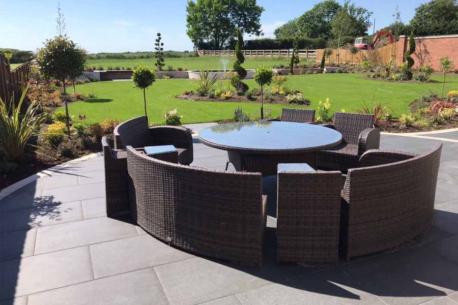 Basalt porcelain patio with a large rattan curved modular dining set, overlooking a large green space with trees and planting.