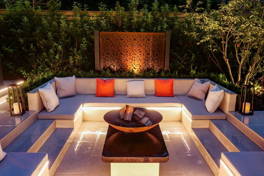 Night time view of Florence Grey Porcelain Paving used around fire-pit in sunken seating area, surrounded by trees.