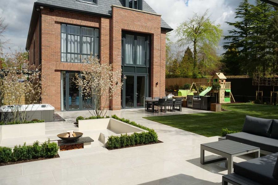 Family garden showing rear of large, modern house. Florence Grey Porcelain Paving used on patio and sunken seating area with fire-pit.