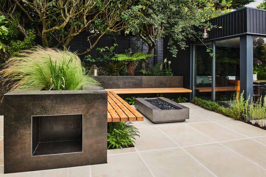 Rectangular firepit on Beige Sawn Sandstone paving, with L-shaped wooden bench cantilevered on raised beds faced with DesignClad.