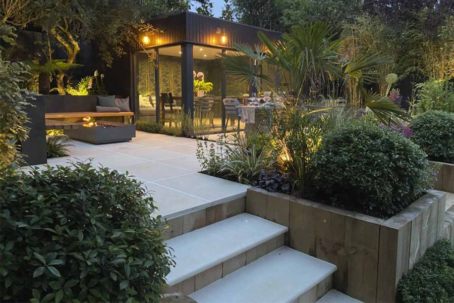 Beige Sawn Sandstone steps rise past raised bed of timber sleepers to patio with illuminated garden room, firepit and bench seating.