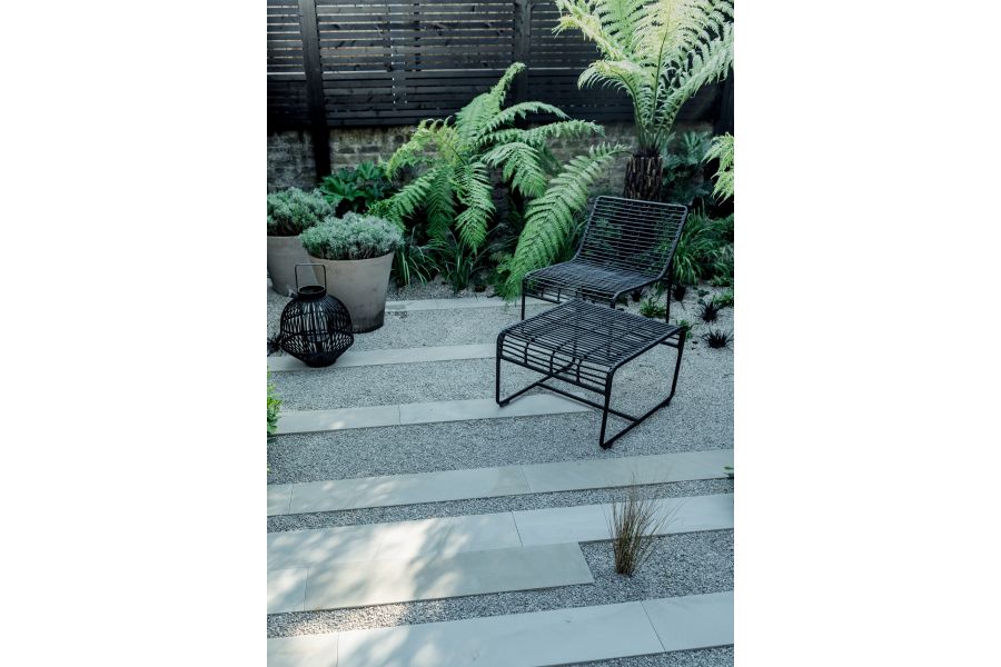 Metal chair and footstool sit on paved area of gravel with page smooth sandstone plank paving. Tree ferns next to boundary wall.