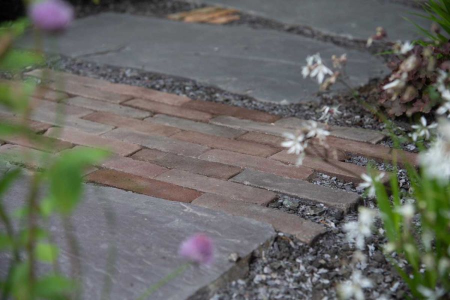 Bergamo Clay Pavers and grey gravel separate natural stone paving slabs in shade seen through blurred flowers. Built by Big Fish.