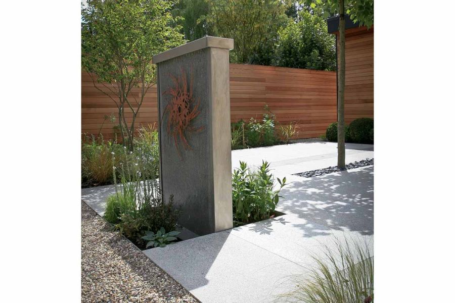 Water wall feature with bog planting on both sides set into Silver Grey granite patio with cedar fence boundary and leafy border.
