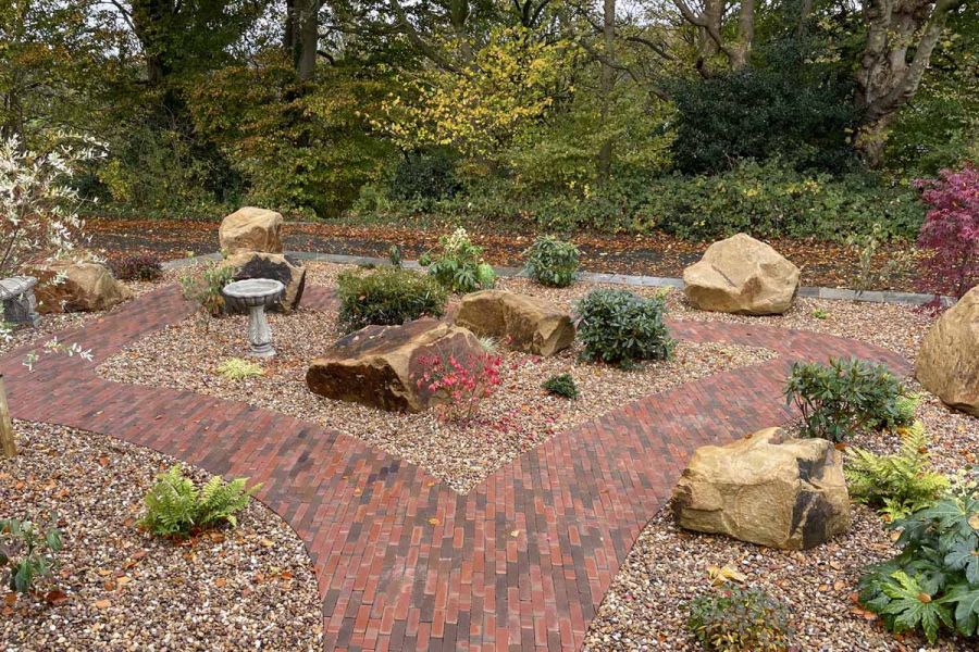 Path of Bexhill clay pavers forks into mirrored curves around boulders, birdbath and small shrubs set into gravel. 