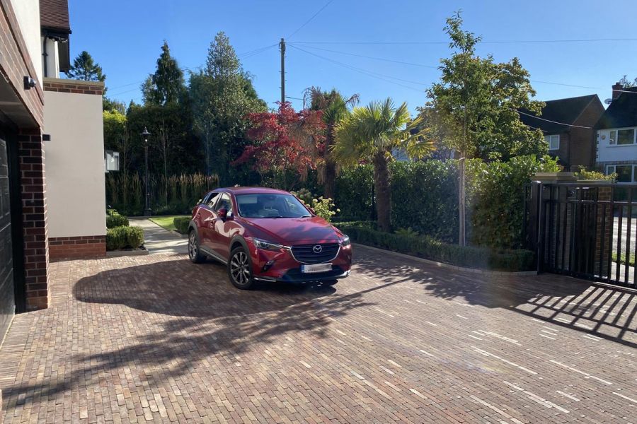 Red Mazda sits on broad driveway across front of house. With gate, hedge and Bexhill clay pavers in running bond pattern.