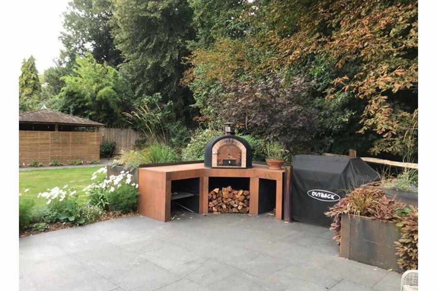 Pizza oven sits on wooden bench in corner of Black granite patio, in front of planted beds, trees and lawn.