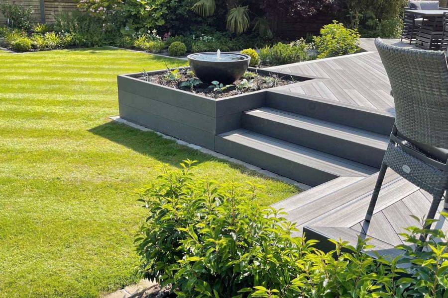 Raised Charcoal composite deck, with bowl water feature on planted bed. 3 steps descend to striped lawn. Design by Blue Tulip.