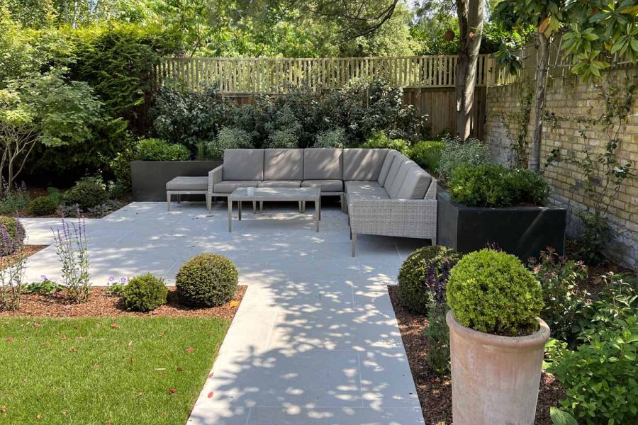 L-shaped outdoor sofa sits with back to raised bed on Florence White porcelain paving square in corner of walled and fenced garden.