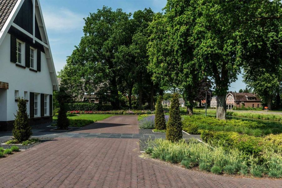 Wide sweep of Bolzano Dutch clay pavers creates flattened U-shape around large planted bed in front of white house.