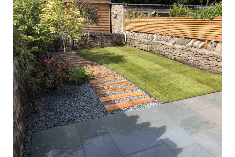 Wood planks set in gravel lead from Brazilian Grey slate patio, past lawn to gate in dry-stone wall, giving entry to garden office.