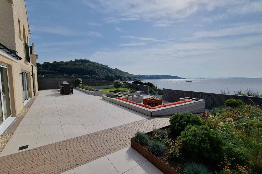 Terraced garden overlooking wide body of water. Strip of Stone Grey clay pavers runs across long light-coloured patio next to house.