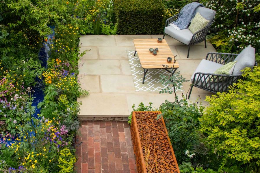 Brick paver path leads by decorative gabion to small square patio of Britannia Buff sawn sandstone paving with 2 chairs and table.