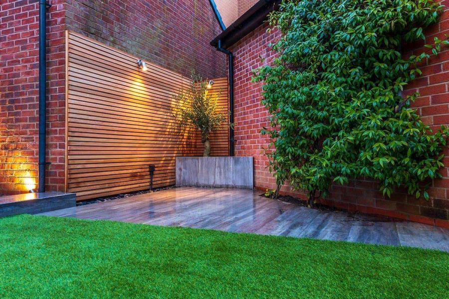 Corner of a small back garden showing an artificial lawn meeting up with a triangular shaped Cinder porcelain patio.