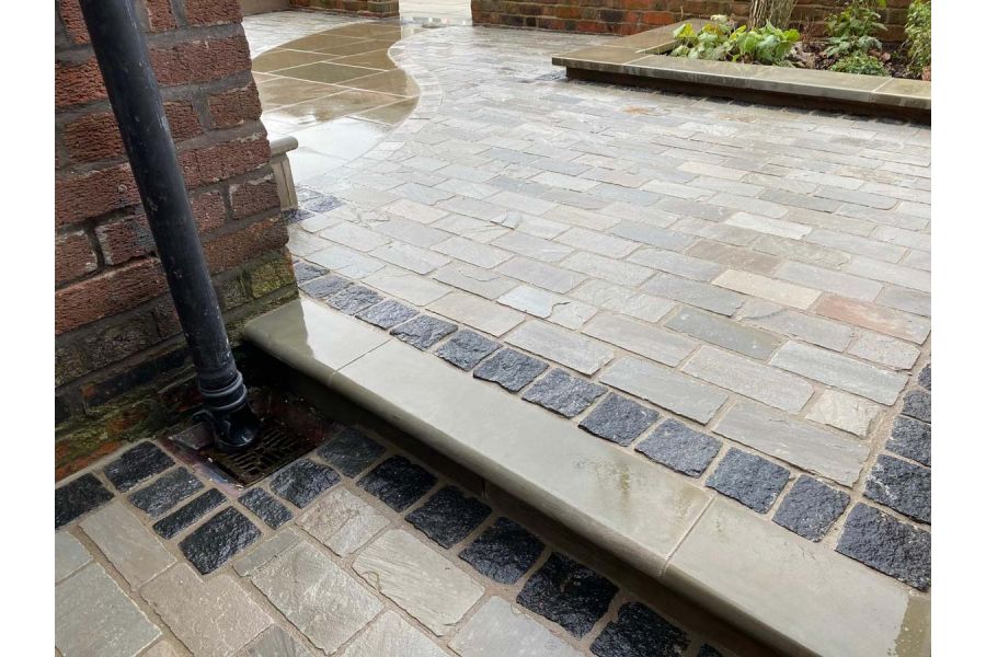 Low view of paved area of Kandla Grey sandstone setts with black granite detailing. Step down by house corner with drainpipe.
