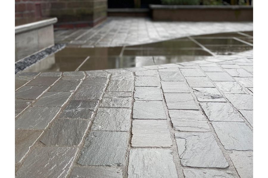 Low view of wet Kandla grey cobble setts with small puddle, showing riven surface. Design by Brockstone Landscape Construction.