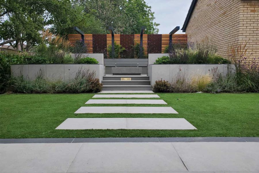 Florence Grey Porcelain paving set into grass lead to steps, flanked by tall raised beds, up to seating area and planting.