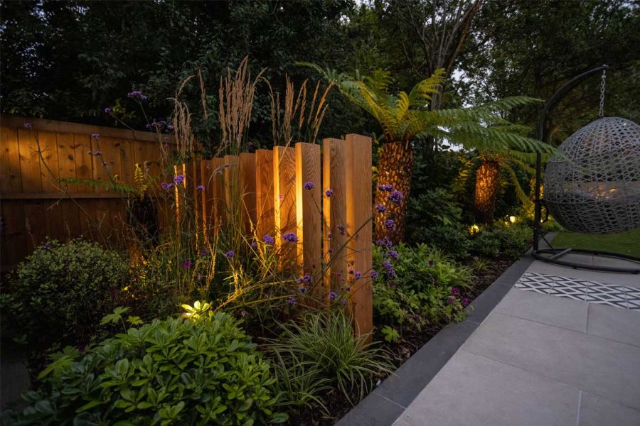 Night view of uplit wooden plank sculpture in deep, planted bed, bordering paved area edged with Platinum Grey Porcelain Planks.