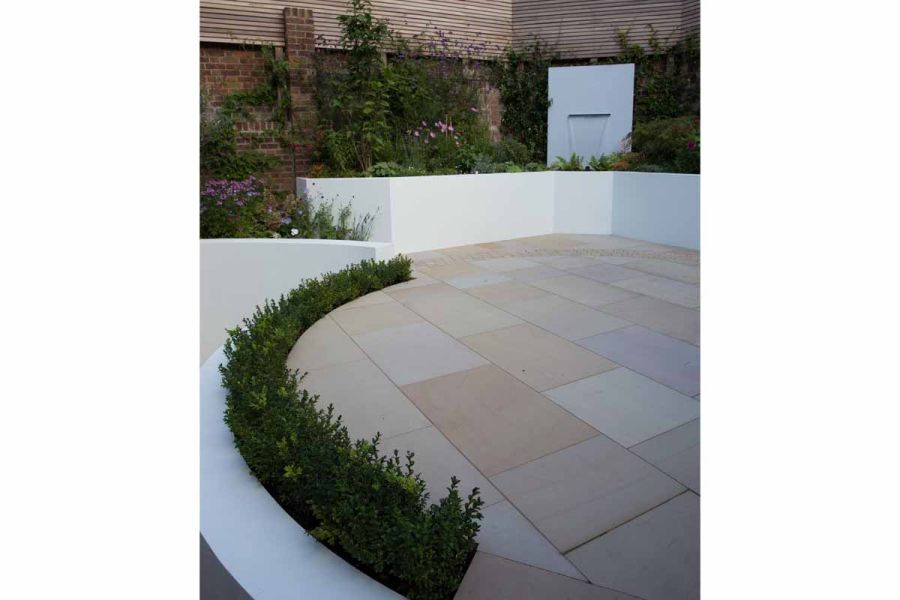 Water feature sits in raised bed on far side of patio of Buff sawn sandstone paving. Slabs are cut to fit a circle outline.