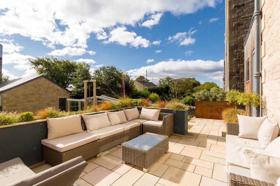 Terrace of mixed sized paving slabs of Buff smooth sandstone, with cream-cushioned furniture and low planting in surrounding wall.