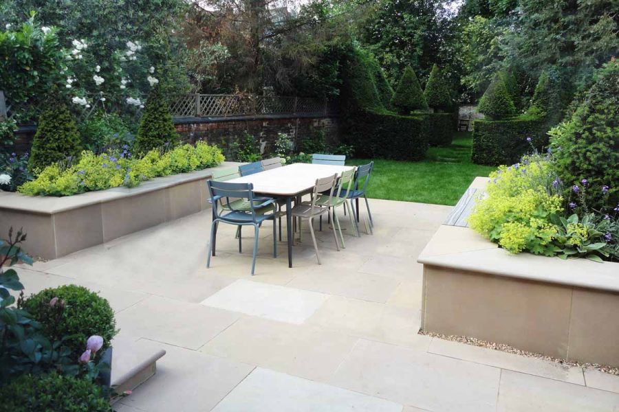 2 large raised beds faced with Buff sandstone smooth paving slabs sit either side of matching paved area with dining set.