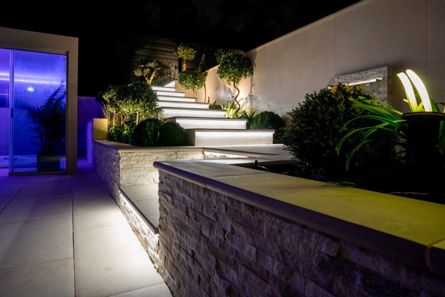 Illuminated flight of Venetian beige bullnose porcelain steps rises from patio alongside high wall. Design by Elitescapes.