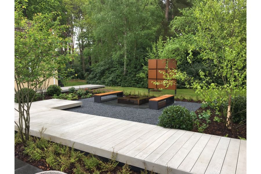 In woodland setting, raised boardwalk in Smoked Oak Millboard decking runs next to seating area with wood-topped benches.