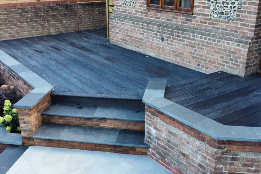 Burnt Cedar Millboard deck with corner cut at angle to allow 2 steps down to paving between brick flanking walls.