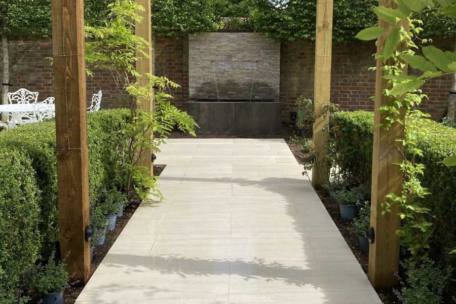 Wide path of Faro porcelain slabs leads between wooden posts and hedges to stone-clad wall with water spout pouring into trough.