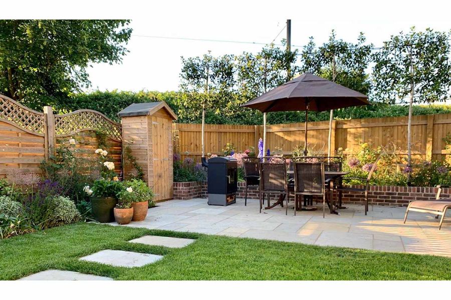 Kota Brown limestone slabs set into lawn lead to matching oblong patio with dining set, tall tool shed and raised bed.