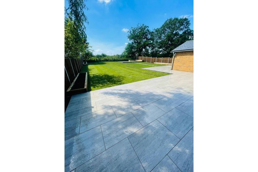View across a large back garden on a summers day.  Patio paved in Travertine Dark Porcelain with dark grout.