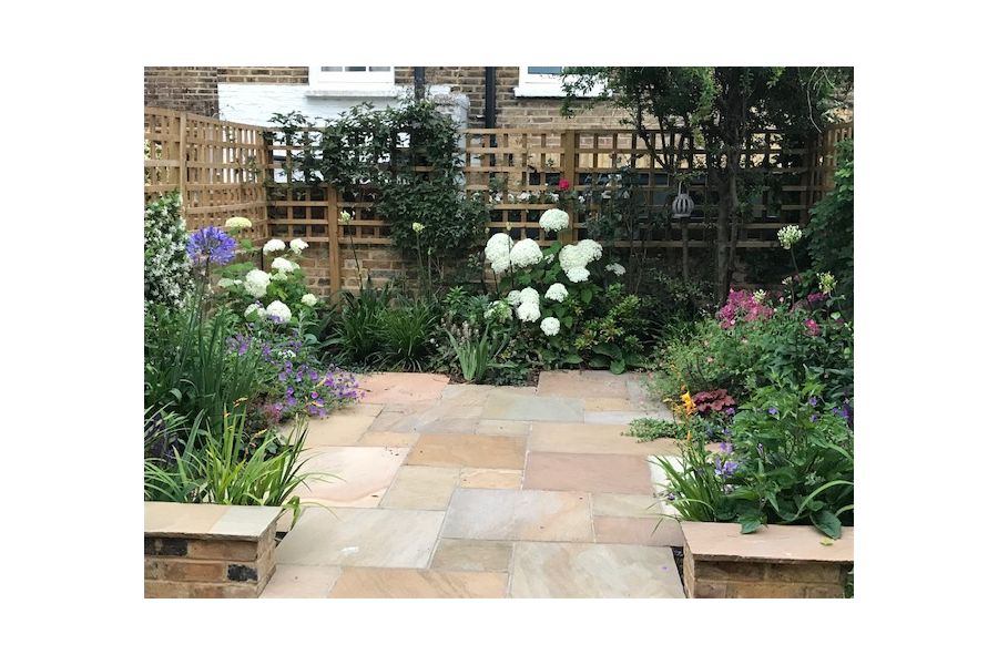 End of narrow urban garden with wall and trellis on 3 sides of Camel Dust Indian sandstone paving edged with planted borders.