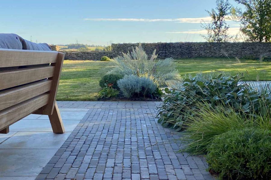 Wide strip of Stone Grey clay pavers edges limestone patio next to planted border and lawn with wall overlooking distant fields.