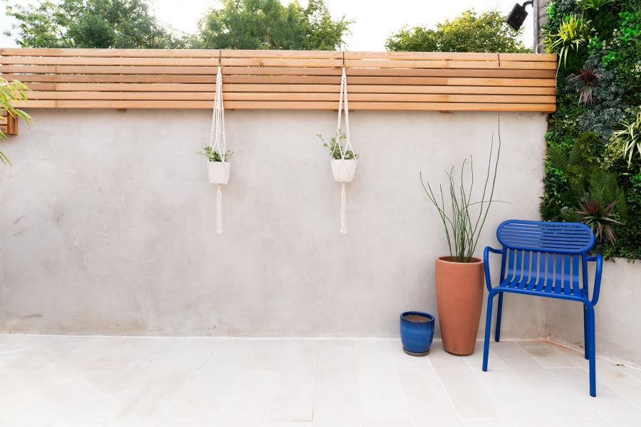 Blue chair and 2 pots on Beige smooth sandstone plank paving next to white wall with plant pots hanging from slatted fence on top.