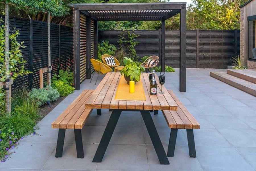 Cement porcelain slab patio with wooden table and benches, chairs under pergola, and slatted fencing.