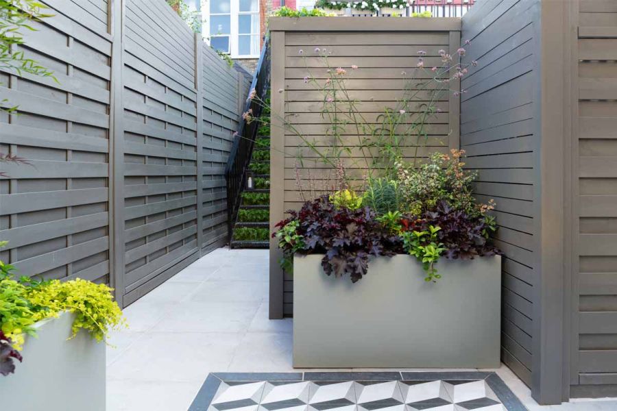 Small back garden with grey slabs, stone-effect, planted through planters and grey wooden fences and metal staircase at the back.
