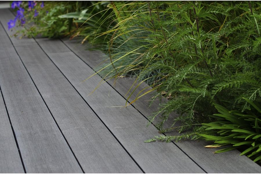 Grasses, long green leaves and blue flowers spill over edge of Charcoal composite decking boards, laid parallel to planted border.