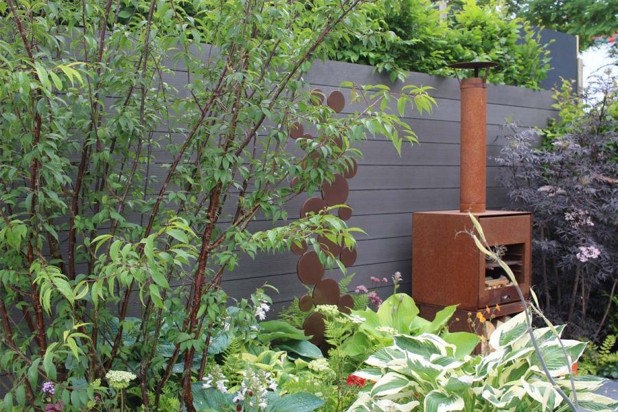 Wall faced with Charcoal DesignBoard decking behind rusty oven with chimney, abstract sculpture and lush planting with hostas.