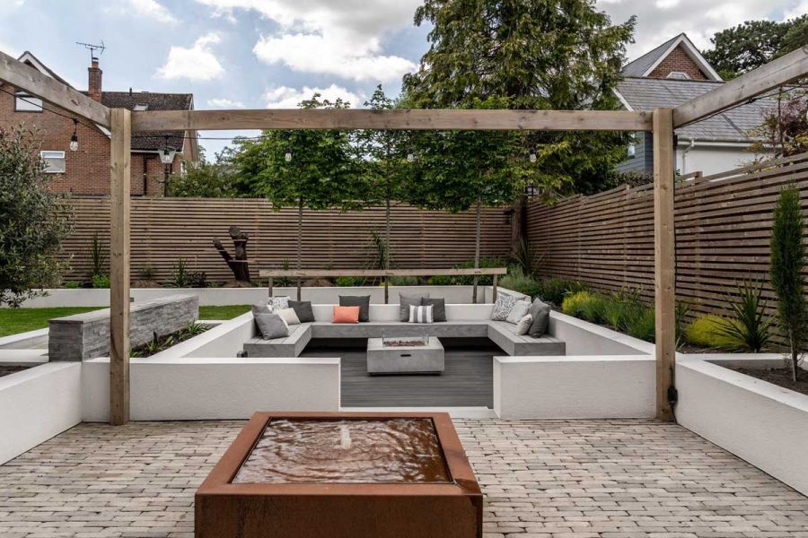 Square seating area enclosed by low walls, set on Charcoal composite decking, behind clay paving with Corten steel raised pond.