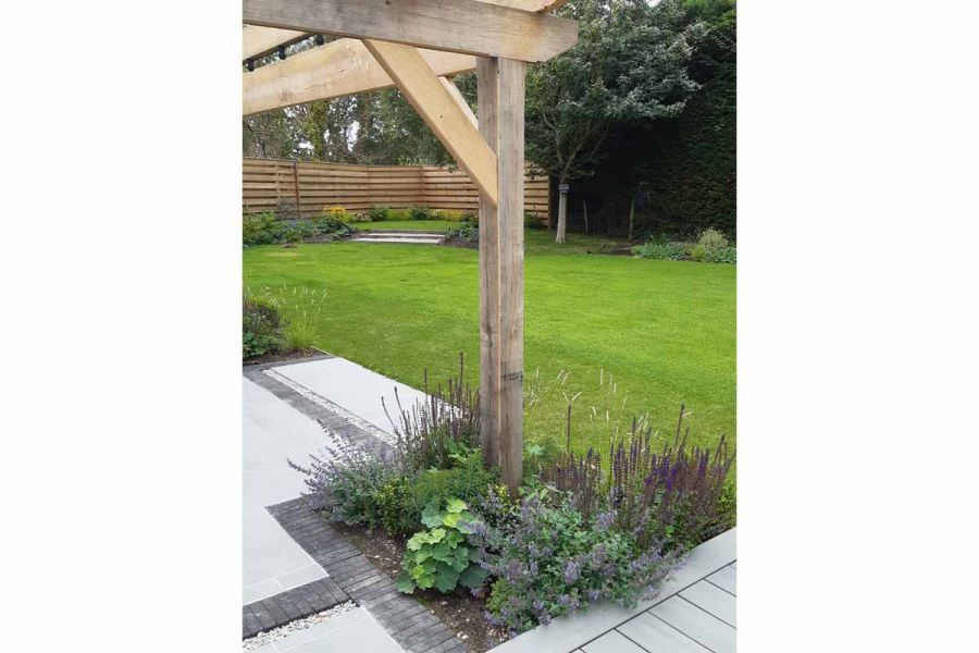 Pale paving and decking with Charcoal Grey clay pavers under wooden pergola next to large lawn by Anna Helps Garden Design.
