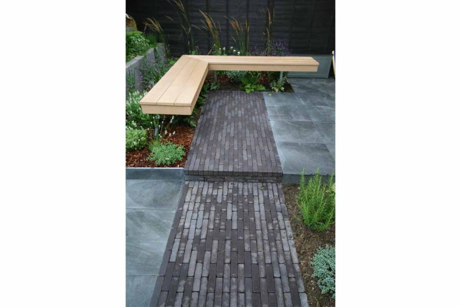 Path of Charcoal Grey garden brick pavers lead to L-shaped wooden bench that sits over planted bed covered with bark mulch.