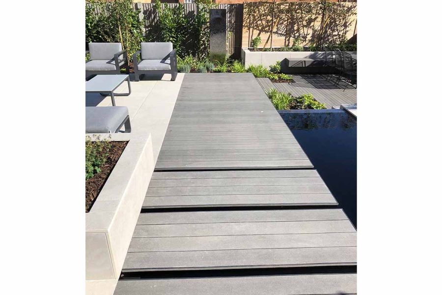 Wide deck of Charcoal composite boards crosses ink-blank rectangular pond next to paved area with outdoor furniture and raised bed.